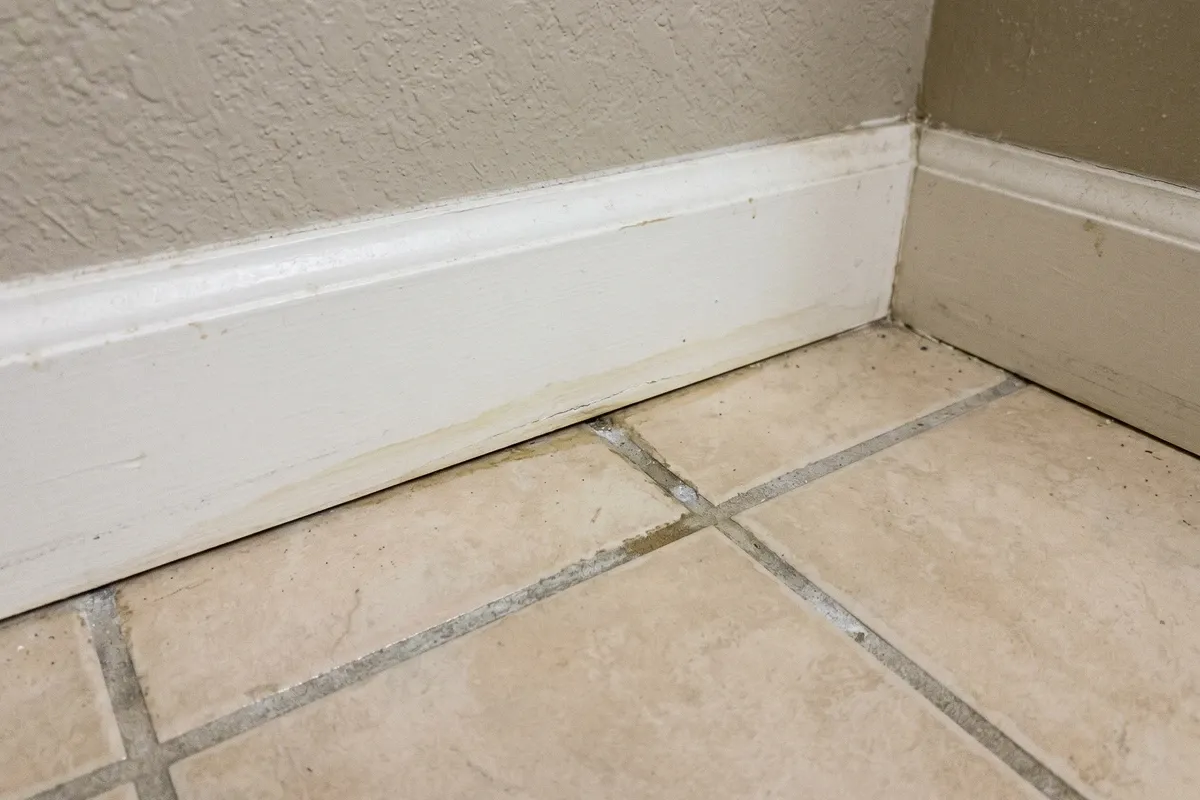 Bathroom baseboard and floor junction in an older Phoenix home showing faint watermark discoloration and slight swelling of baseboard trim from a slow undetected leak, aged beige tile, ordinary bathroom lighting.