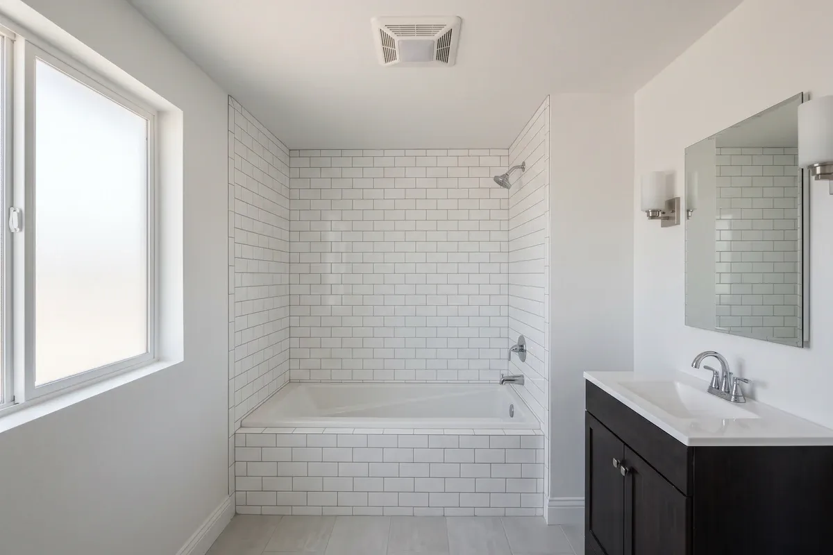 New construction bathroom interior with white tile, a bath exhaust fan grille centered on the ceiling, clean new fixtures, soft window light.