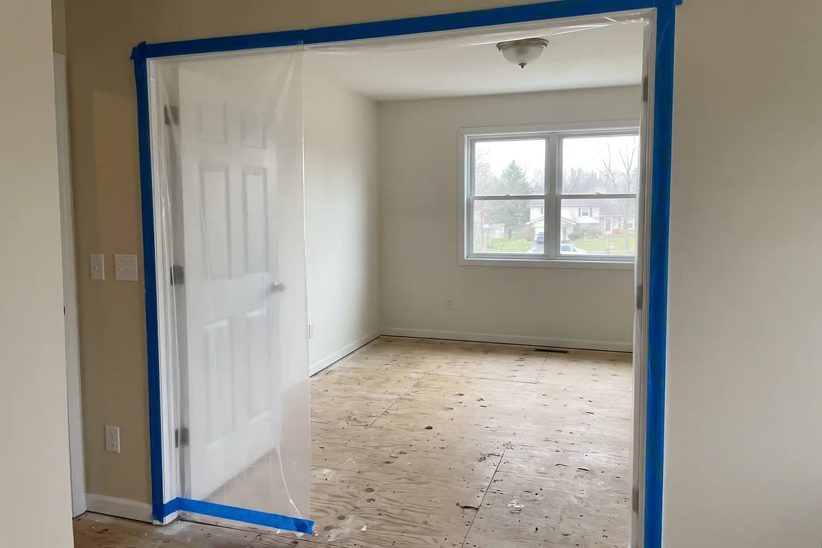 An emptied bedroom with clear plastic sheeting taped across the doorway from floor to ceiling, bare subfloor, daylight through an uncovered window