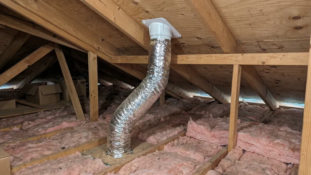 Silver insulated flex duct running from attic floor up toward a white plastic boot fitting at the roof deck, proper residential attic installation with fiberglass insulation below, diffuse natural light.