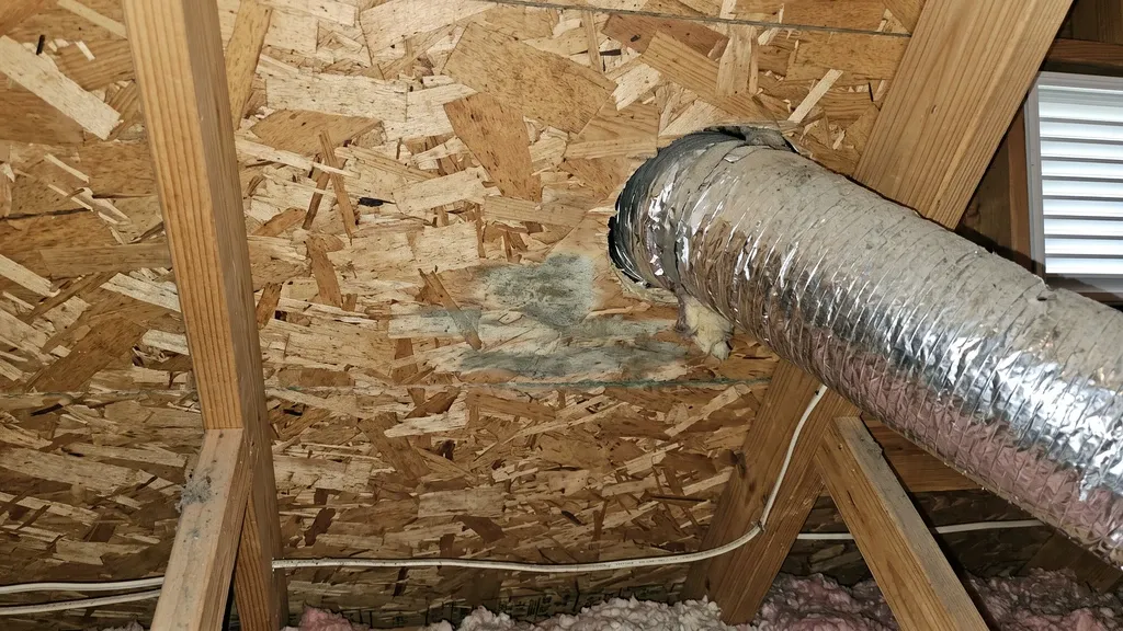 Small patch of gray-green mold discoloration on raw OSB plywood near a duct penetration on an attic roof deck, modest early-stage staining visible under diffuse attic light.