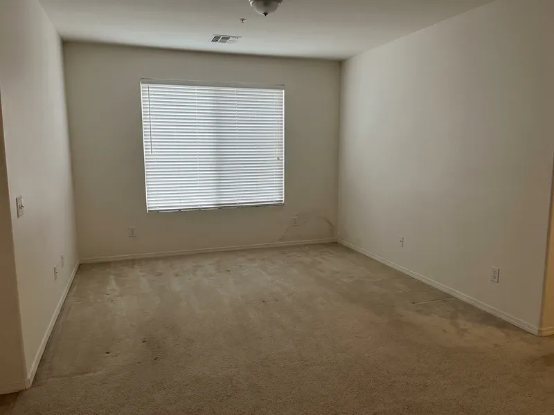 The interior of an empty Phoenix home living room with closed blinds and a faint water stain visible on the lower wall near the baseboard, the room vacant and still.