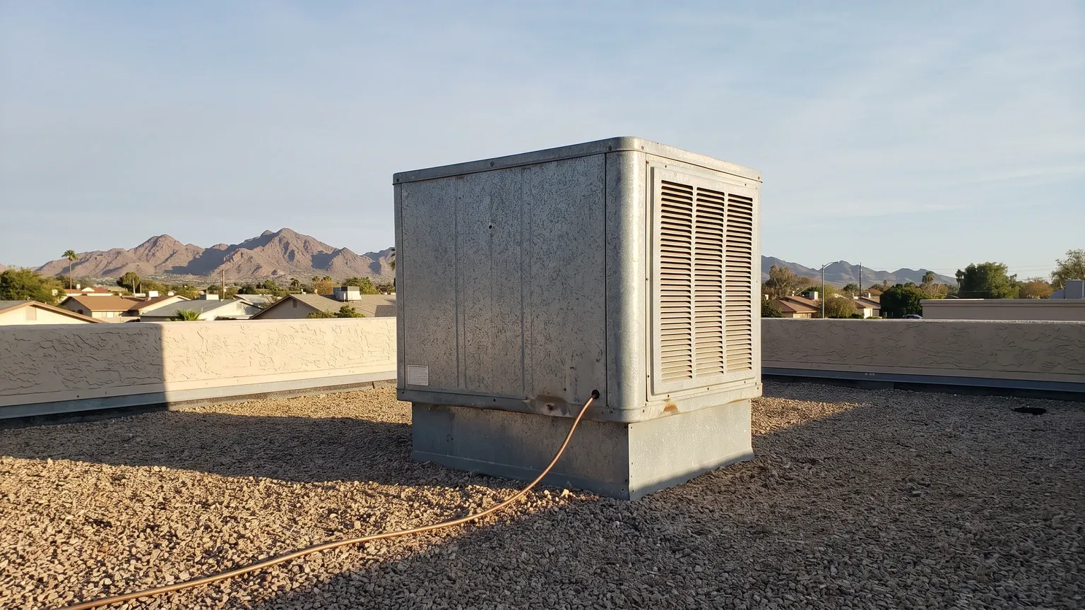 A boxy galvanized sheet-metal evaporative swamp cooler on the low flat roof of a single-story Phoenix stucco home in the low autumn sun, dusty weathered panels and a visible water supply line, desert mountains on the horizon.