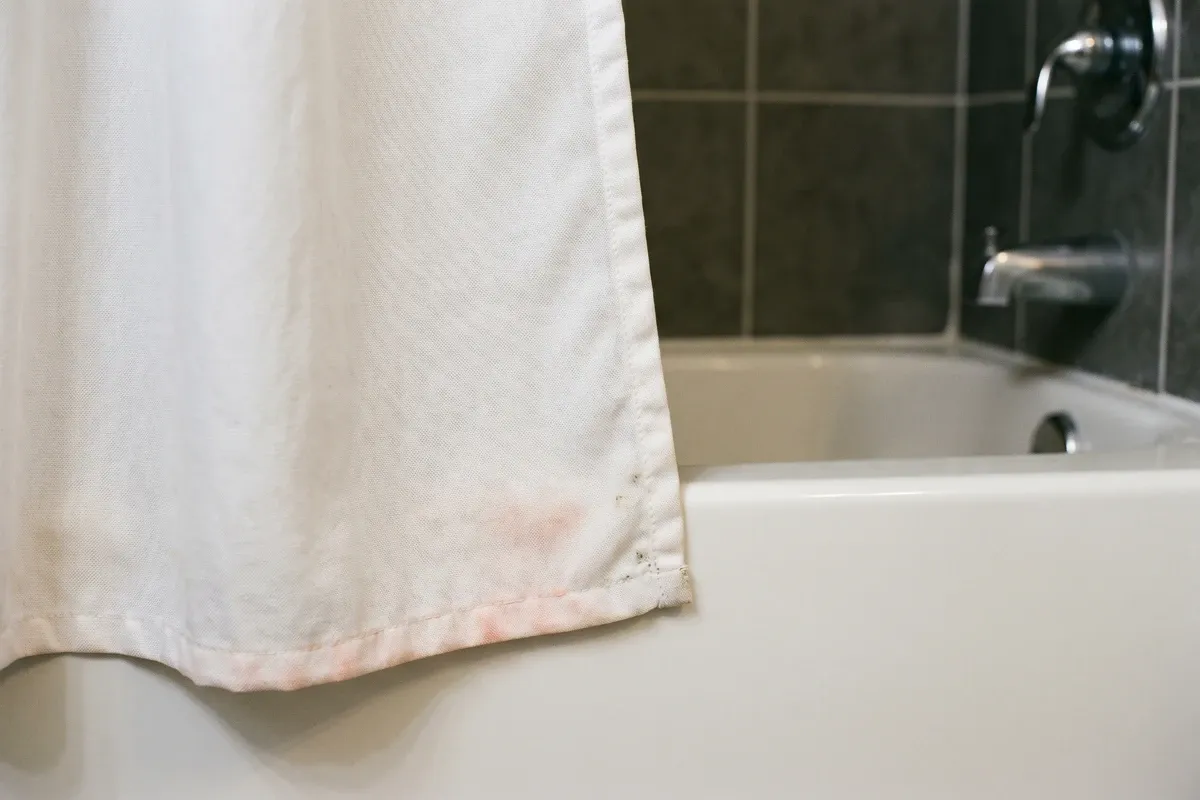 Shower curtain corner showing mild pink-orange discoloration and a small amount of mildew, photographed close up in warm bathroom light.