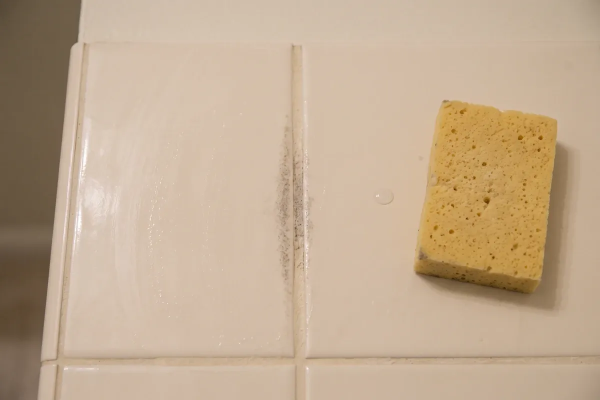 Close-up of glazed bathroom tile and a grout line with light early staining, a sponge nearby