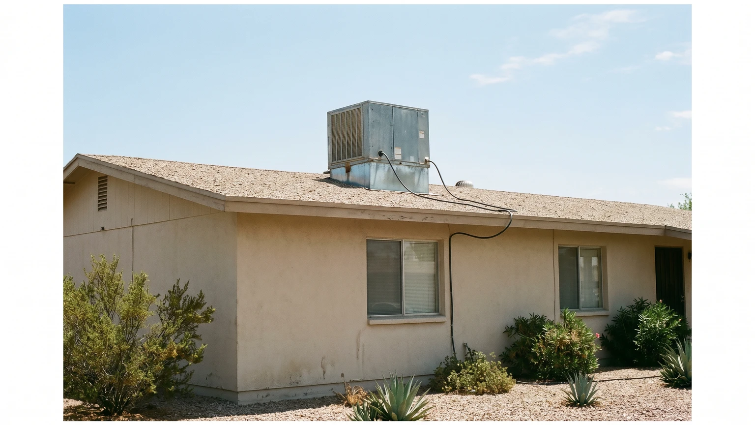 A galvanized sheet-metal evaporative swamp cooler on the flat roof of a single-story Phoenix stucco home in bright midday desert sun.